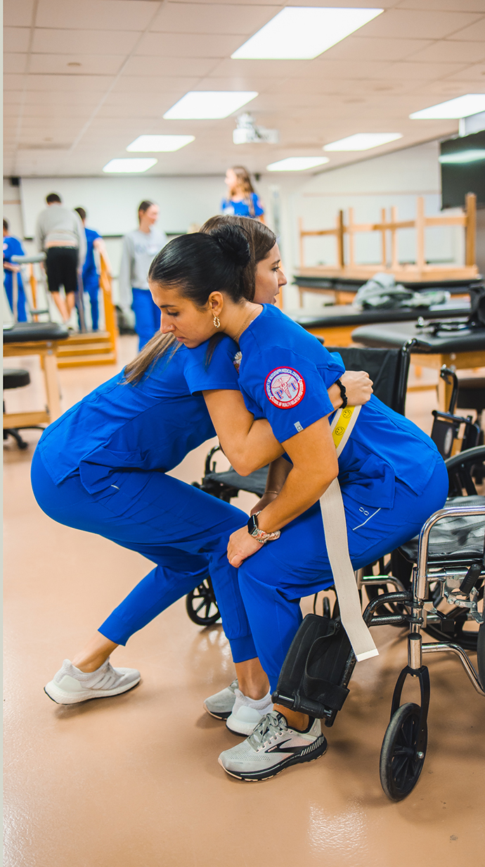A young woman wearing blue medical scrubs lifts another young women froma wheelchair.