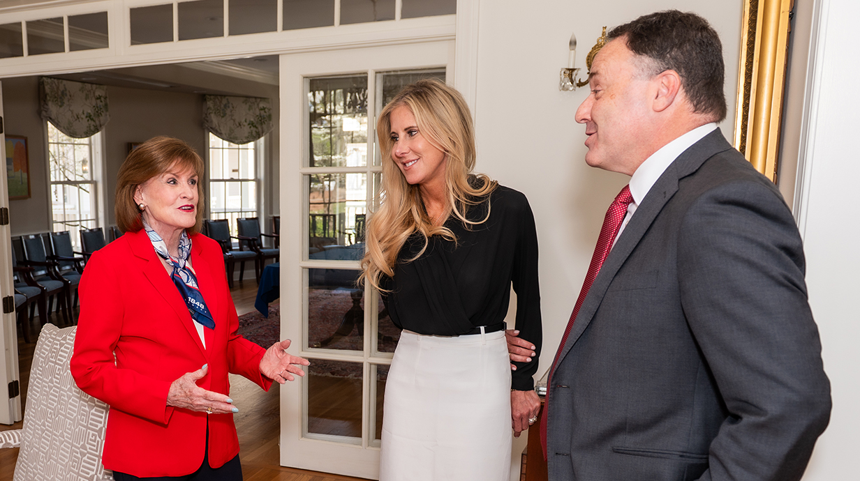 An older woman wearing a red jacket chats with a woman and a man wearing a dark suit.