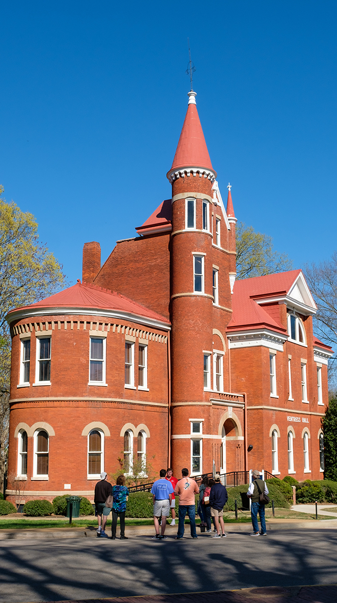 A gothic red brick building with round turrets on top sits on the edge of a wooded park.