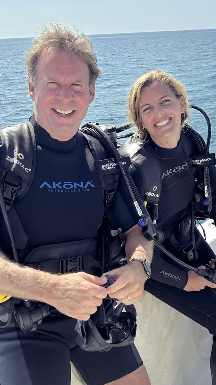 A man and a young woman wear scuba gear while sitting on the side of a boat.
