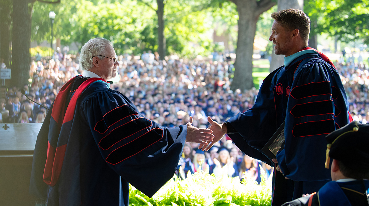 Two men wearing graduation robes shake hands on an outdoor stage in front of a huge crowd of people.