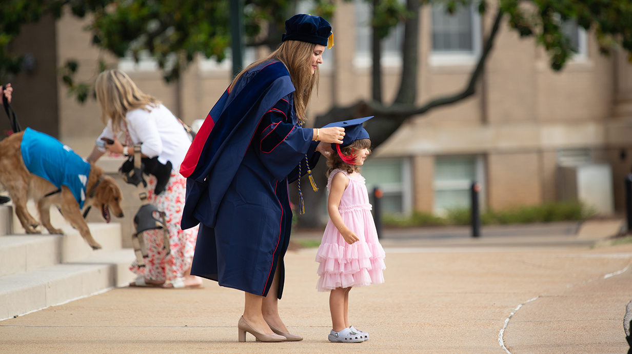A woman wearing graduation robes put her cap on a young girl's head in front of a large building.