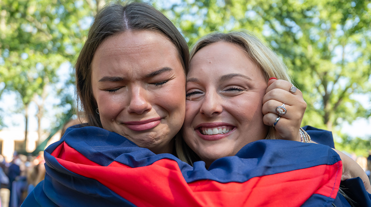 Two young women wearing graduation robes share a hug.