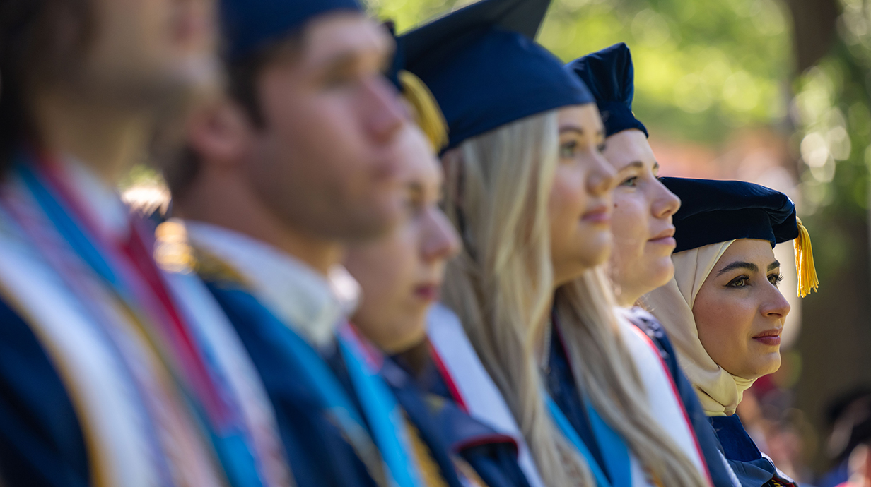 A line of young women wearing graduation robes listen to a speaker.