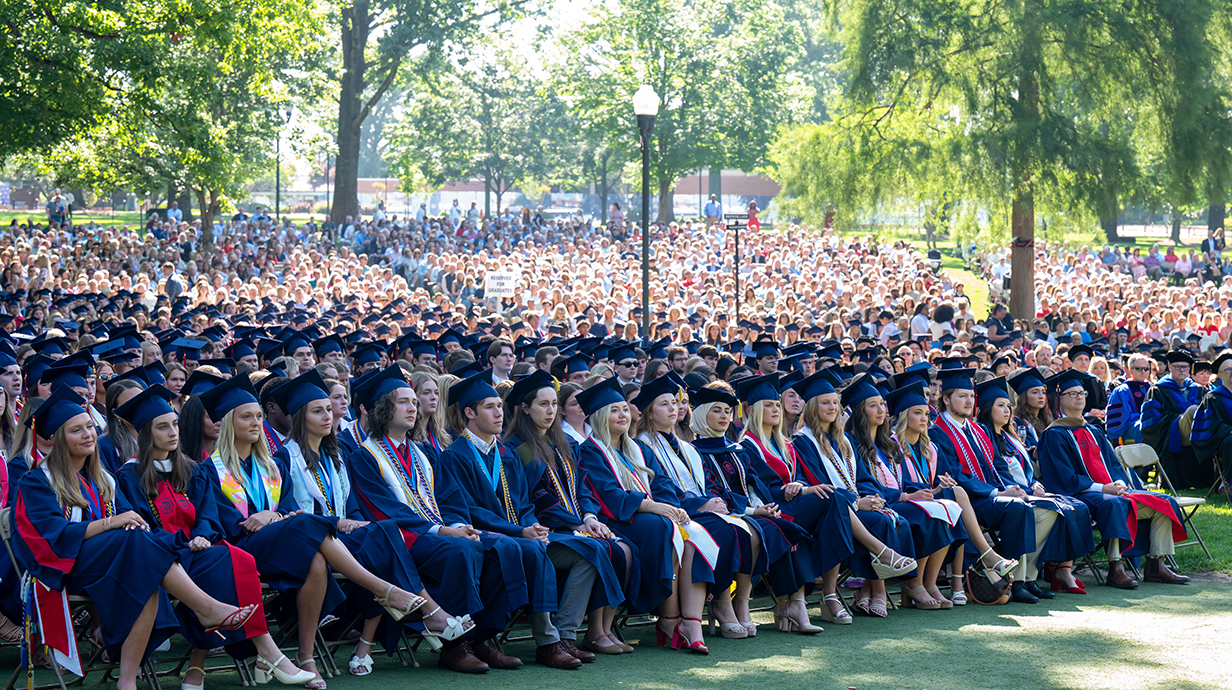 A large group of people sit in chairs in a tree-lined park for graduation.