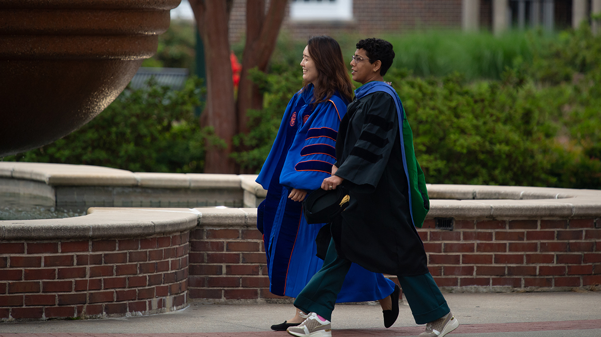 A man and a woman, both wearing graduation robes, walk along a sidewalk in front of a stone fountain.