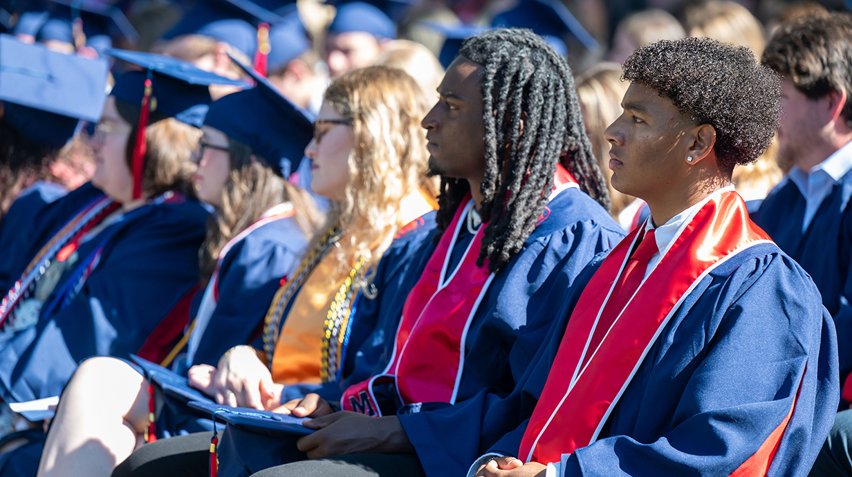Several young men sit in a graduation ceremony on a sunny day.