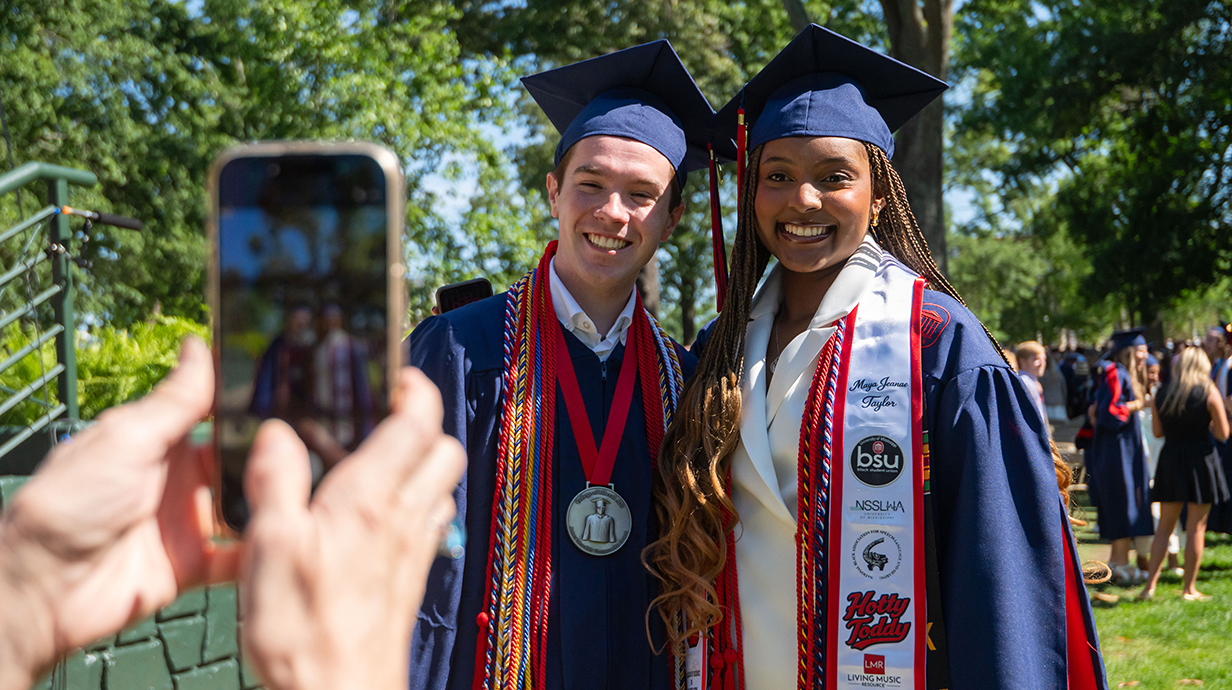 A young man and young woman wearing graduation robes pose for a photo.
