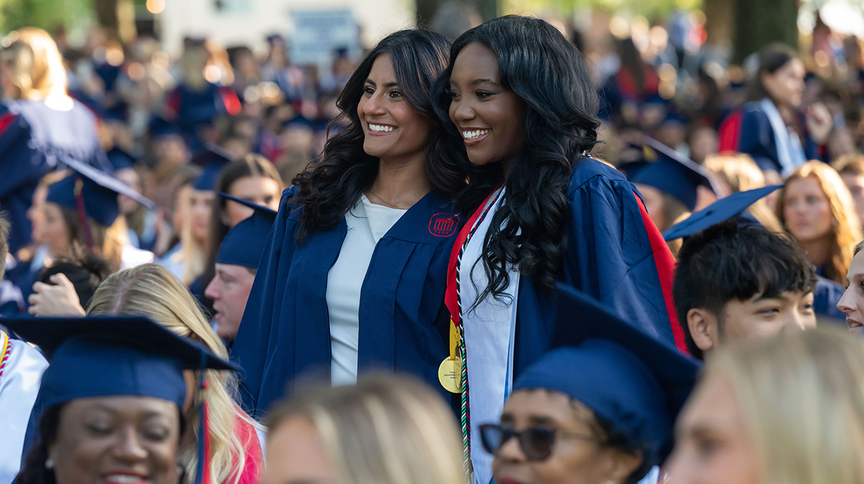 Two young women wearing graduation robes stand in a crowd of people seated outdoors.
