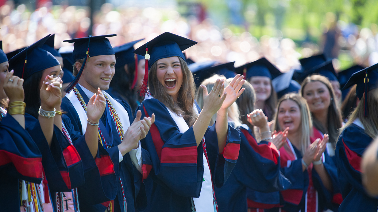 A group of young women stand among a crowd of people wearing graduation robes.