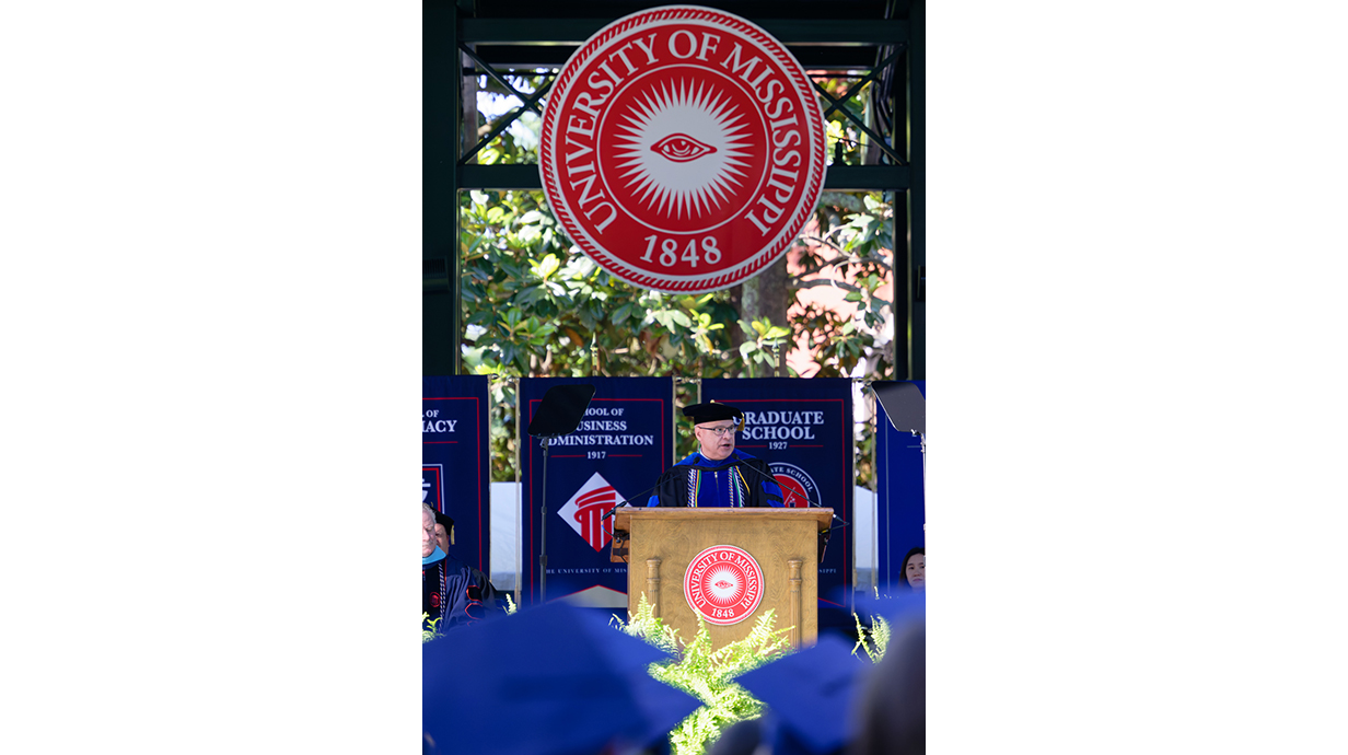 A man speaks on a stage under a large red logo of a university seal.