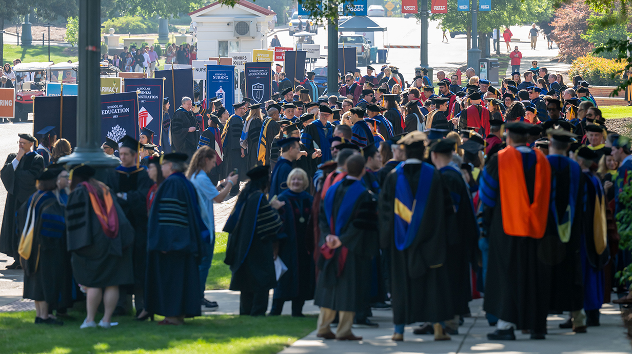 A large group of people wearing academic regalia stand on a sidewalk outside a park.