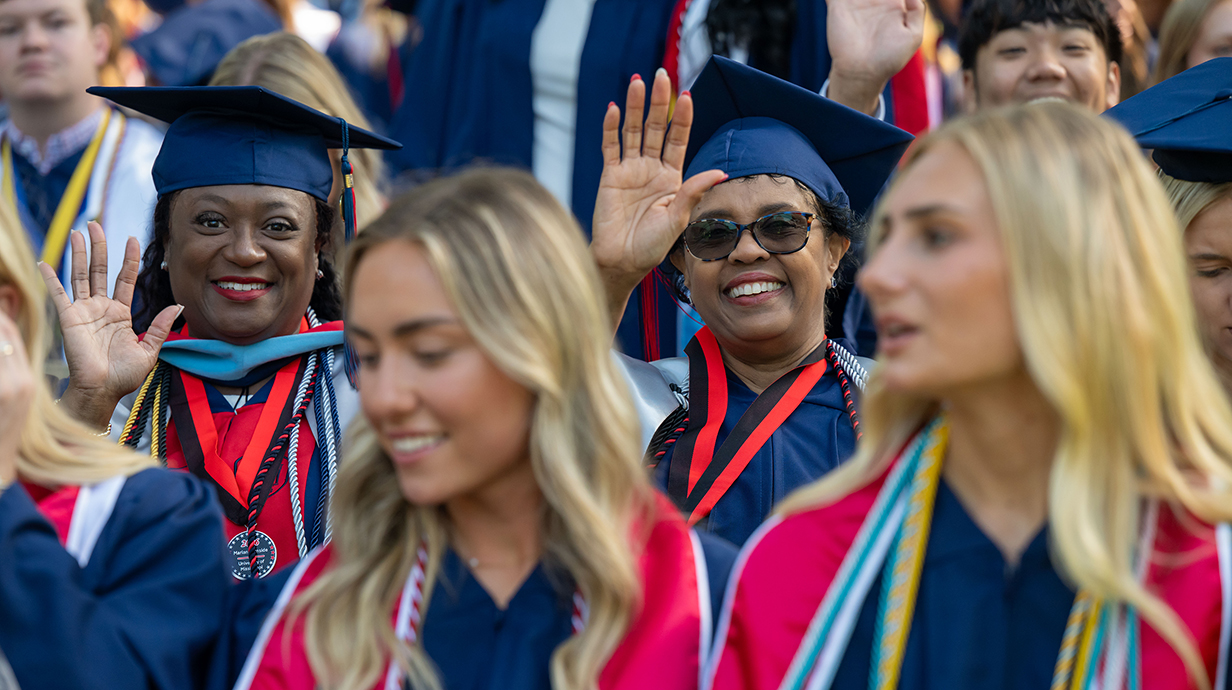 Two women wearing graduation robes wave to the camera while seated among a crowd.