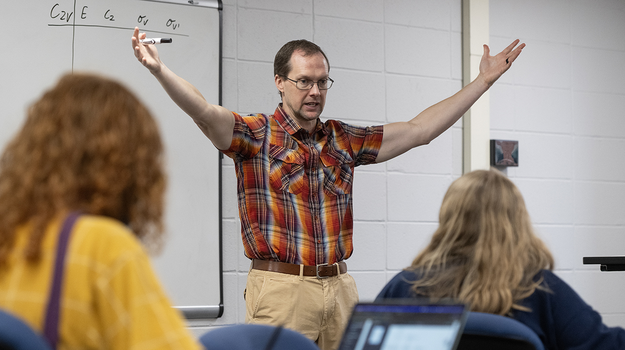A man wearing a plaid shirt stands with his arms raised in front of a whiteboard at the  front of a classroom.