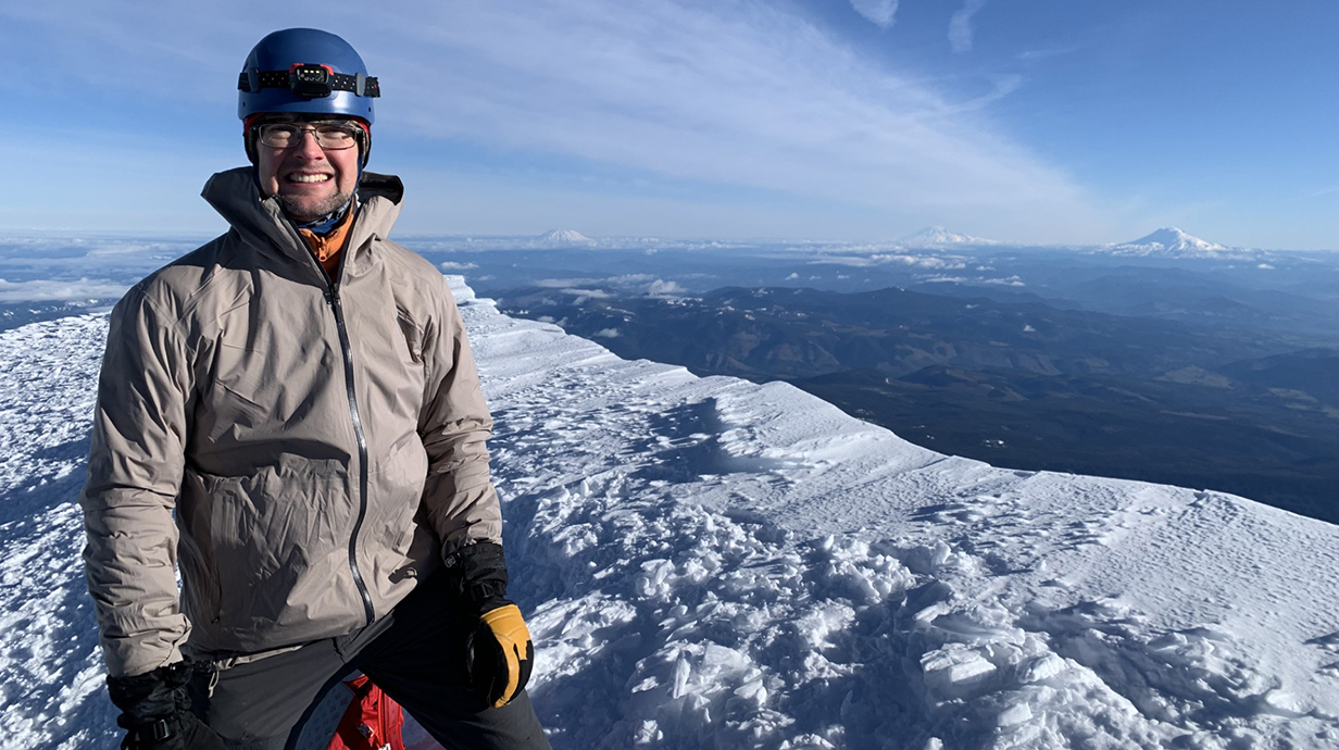 A man stands on a snow-covered mountain with other peaks in the background.