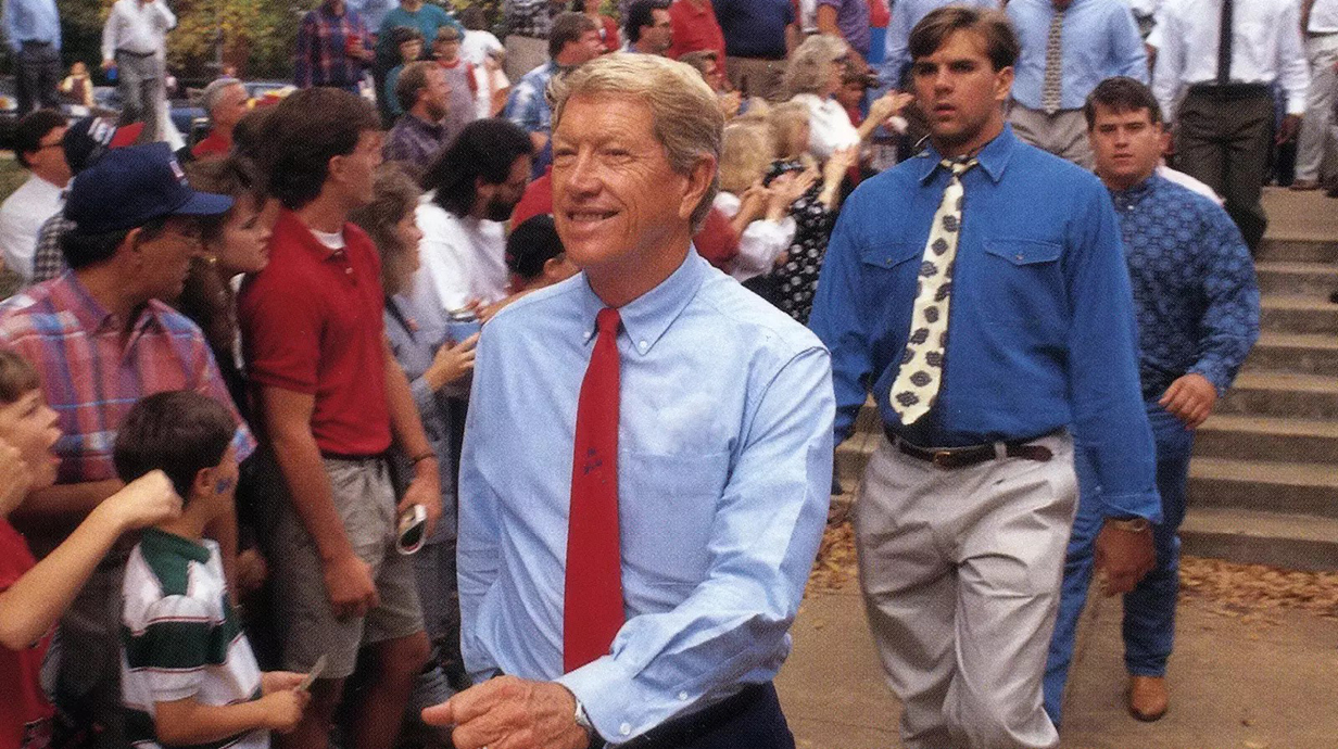 A man wearing a dress shirt and tie leads a group of young men as they walk past a crowd of people lining a sidewalk.