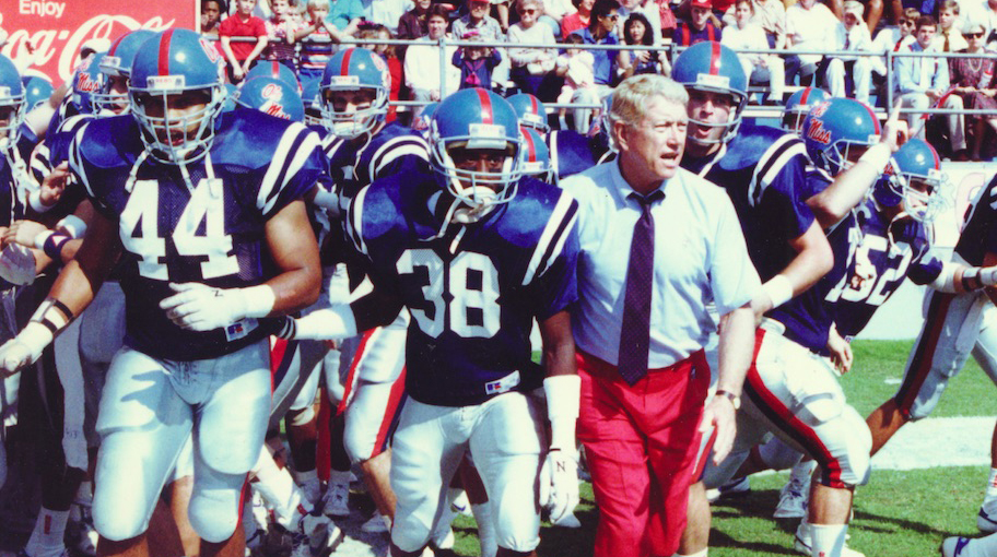 A man wearing a dress shirt and tie leads a football team onto the field.