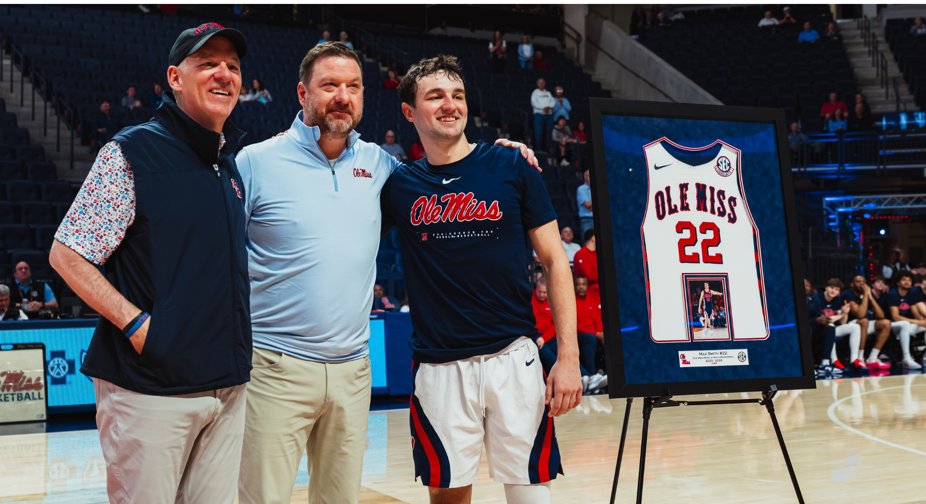 An image of Scott Smith, Coach Chris Beard, and Max Smith next to a framed jersey at Max's graduation recognition ahead of a basketball game.