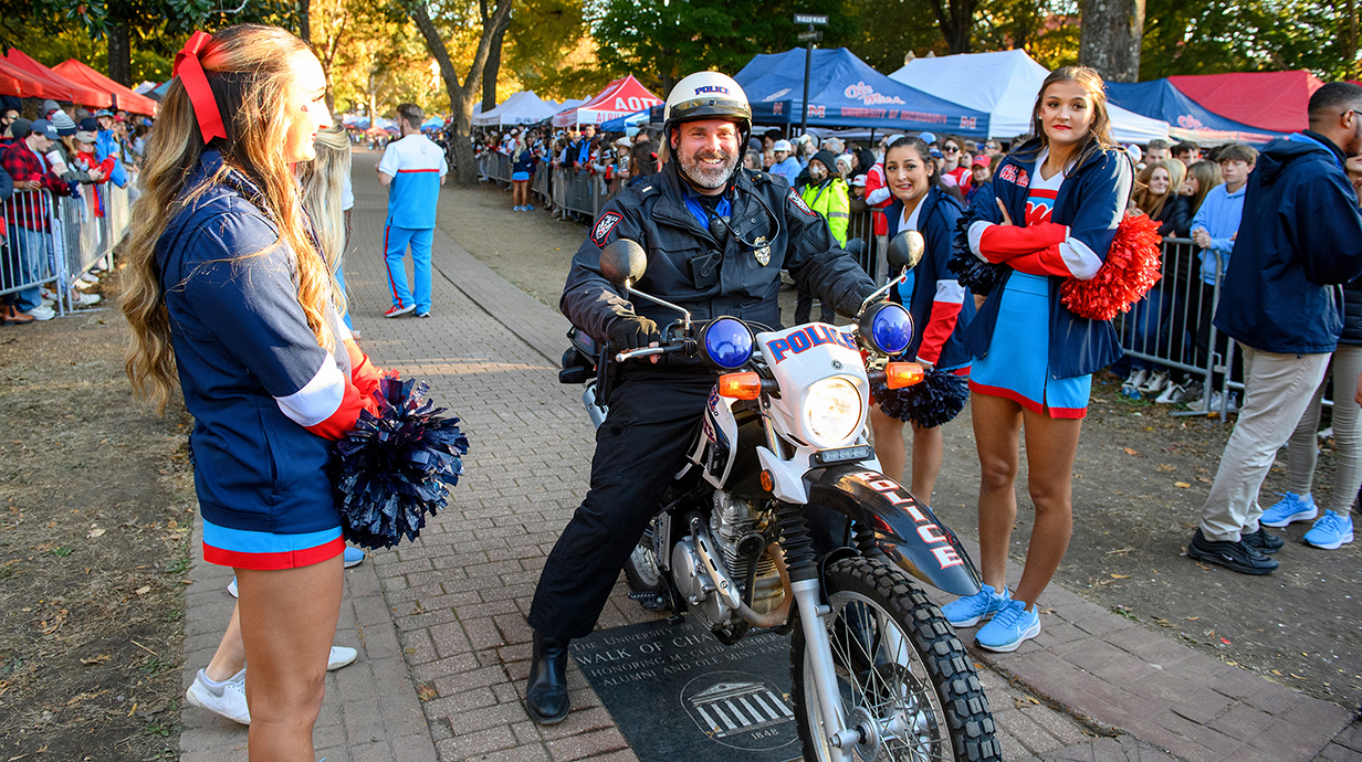 A police officer sits astride a motorcycle on a walkway through an outdoor park lined with people and cheerleaders.