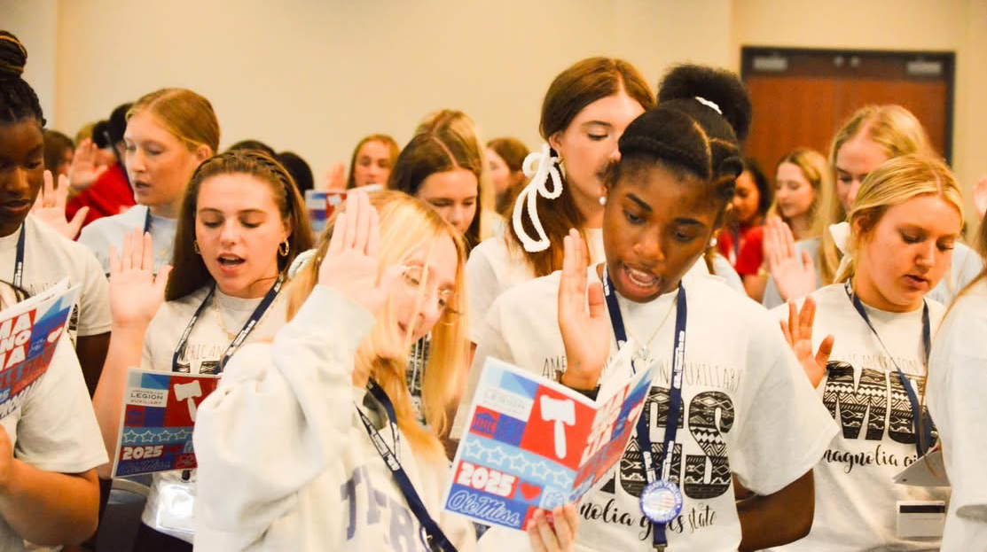 A large group of young women raise their hands as they recite a pledge in a meeting room.