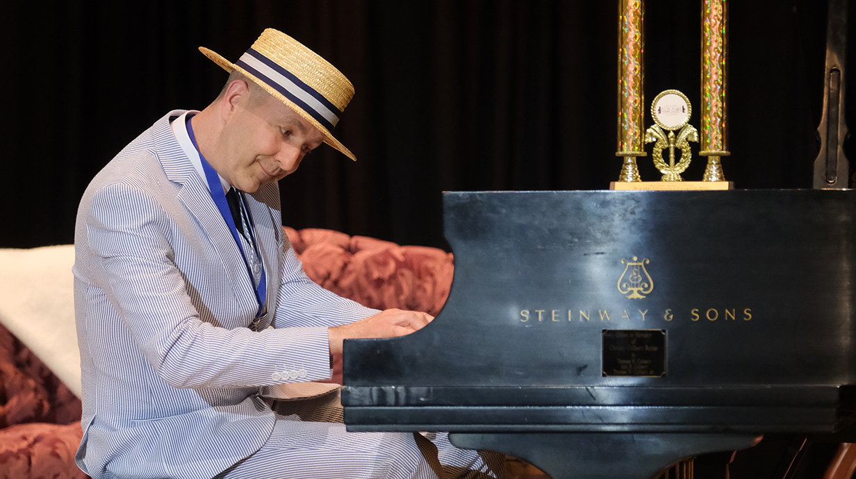 A man wearing a seersucker jacket and a boater hat plays a piano with a trophy sitting on top of it.