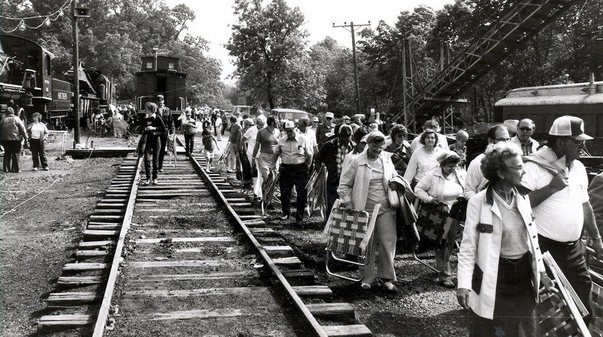 A large group of people carrying lawn chairs walk along railroad tracks.