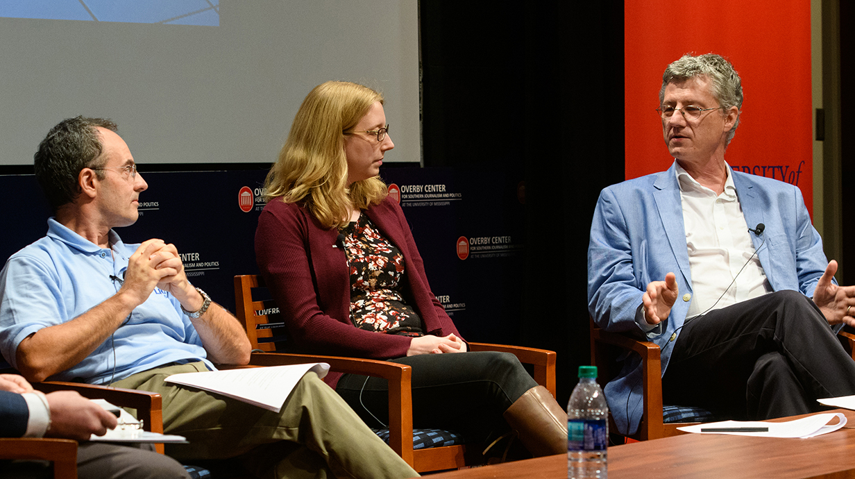 Two men and a woman sit in chairs on a stage backed by a red-and-blue banner.