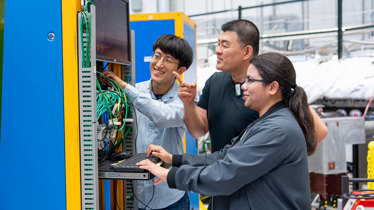 Two men and a woman work at a computer terminal attached to a large piece of electronic equipment.