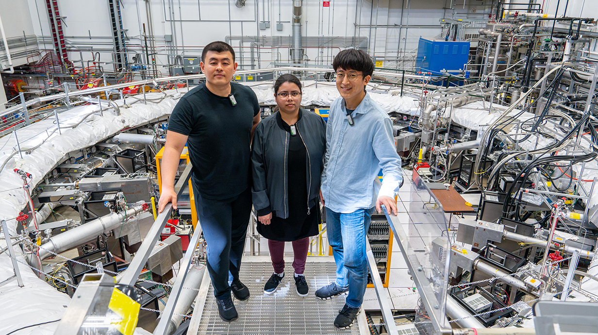 Two men and a woman stand on a platform overlooking a giant laboratory lined with electronic equipment.