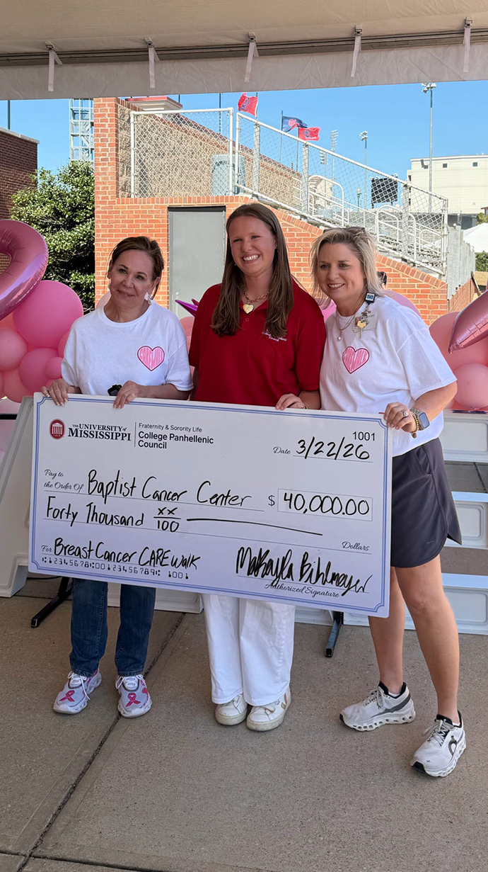 Three women hold a giannt check in front of banners and balloons.