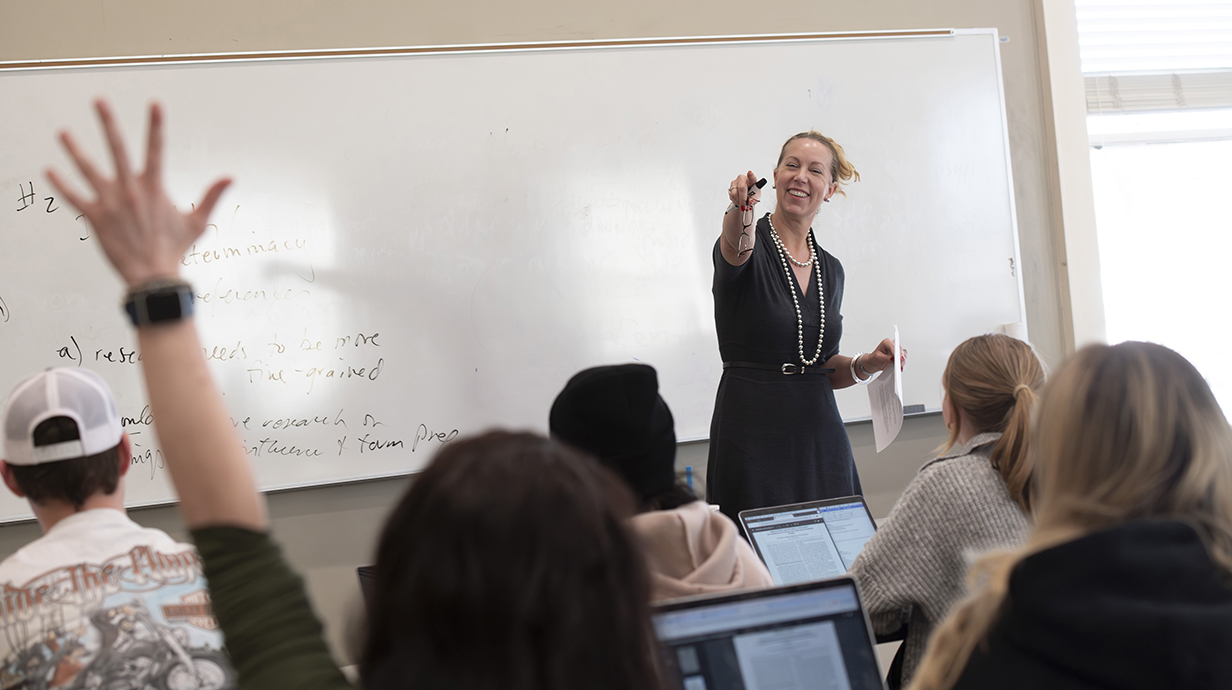 A woman standing at the front of a classroom points to a student's upraised hand.