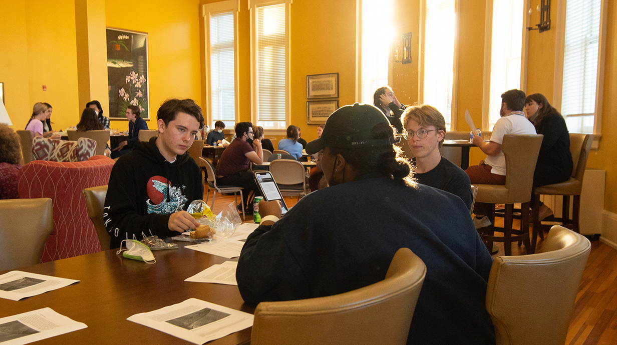 A group of young people sit around a table in a room lined with tall windows.