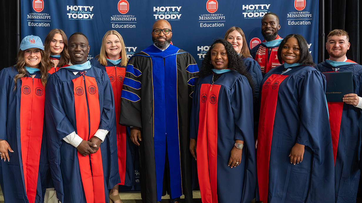 A group of men and women, all wearing graduation robes, stand in front of a dark blue backdrop.