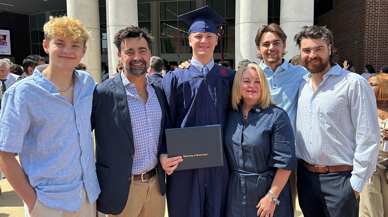 A young man wearing a graduation robe and holding a diploma folder stands outside, surrounded by several family members.
