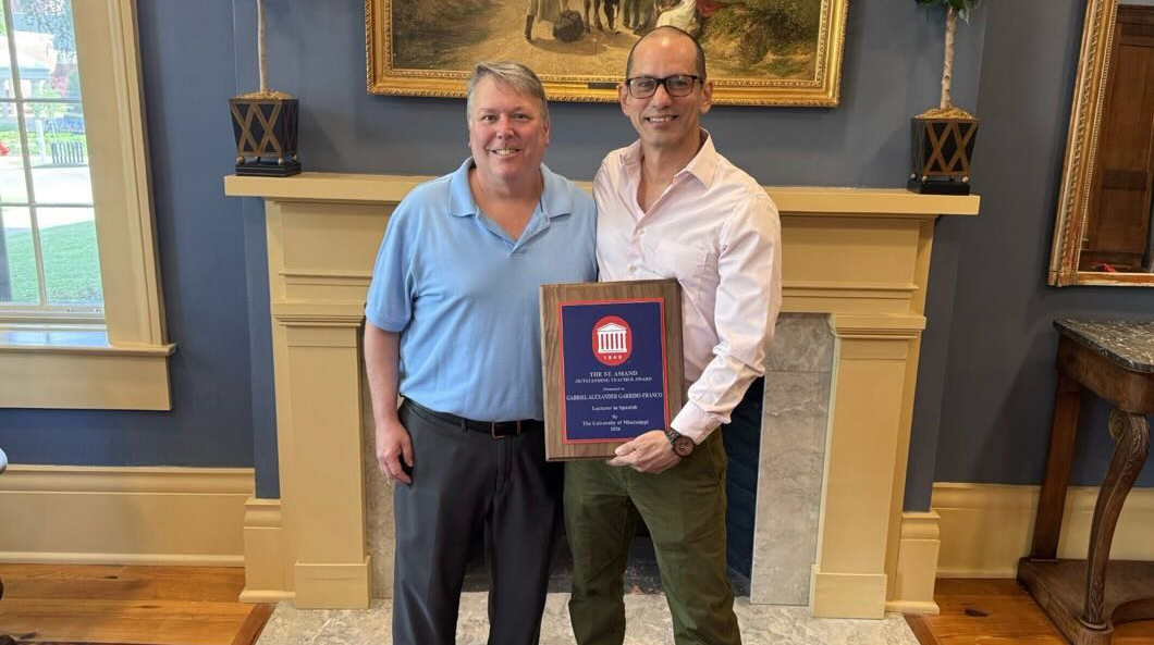 Two men hold an award plaque in an ornate parlor.