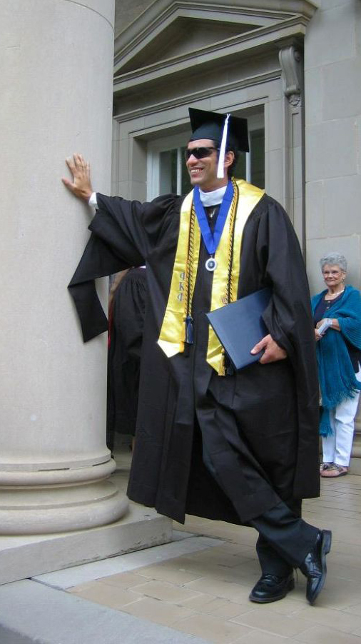 A young man wearing graduation robes leans against a large stone column.