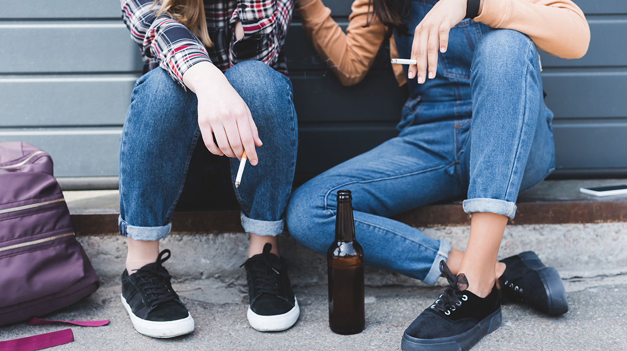 Two young people holding cigarettes sit on a curb with a beer bottle sitting between them.