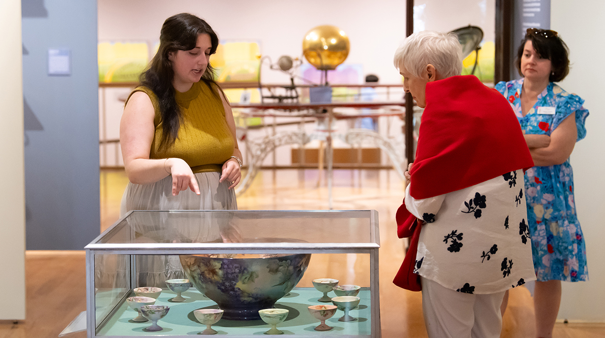 A woman talks with an older woman as they stand near a display case of ancient pottery.