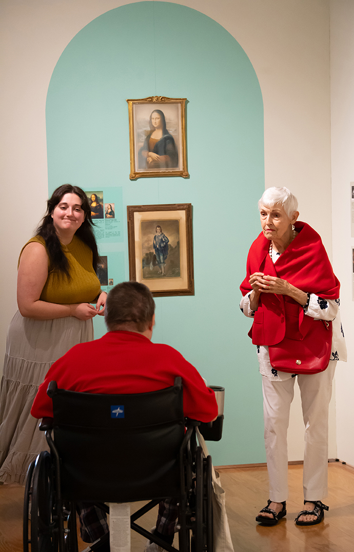 A woman talks to an older woman and a man seated in a wheelchair in an art gallery.