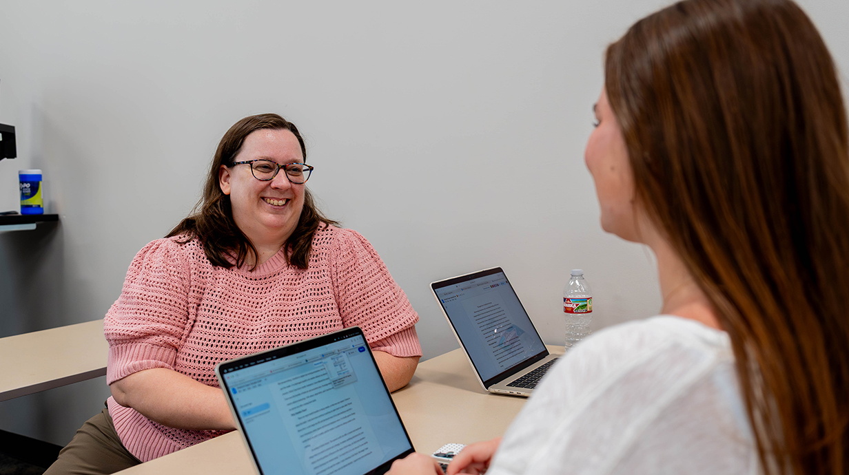 A woman sits on one side of a narrow table while talking to a young woman with a laptop computer on the opposite side.