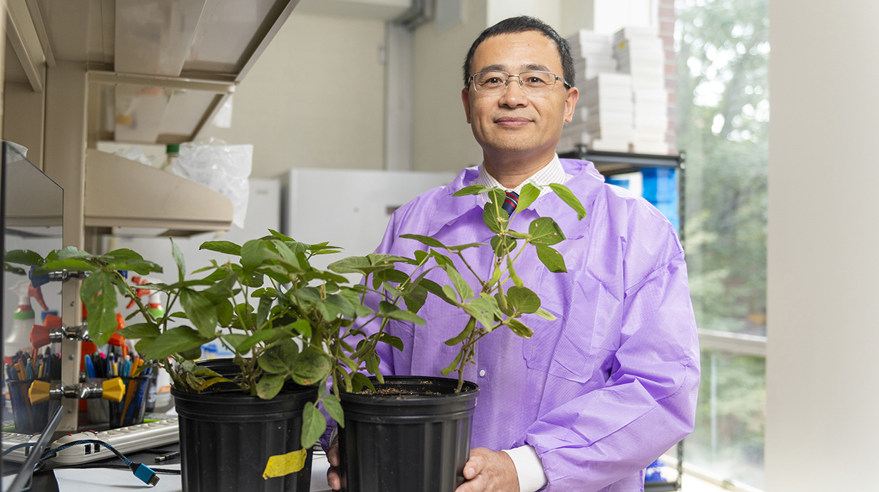 A man wearing a lab coat stands with a potted plant in a laboratory.