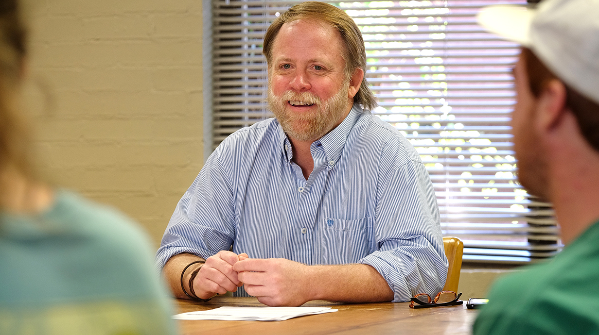 A man seated at the end of a table smiles and listens to young people seated around it.