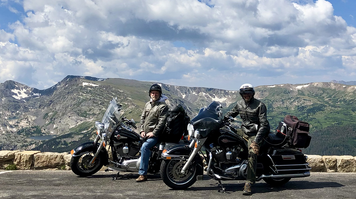 Two men sit on motorcycles at an overlook in front of a mountain range.