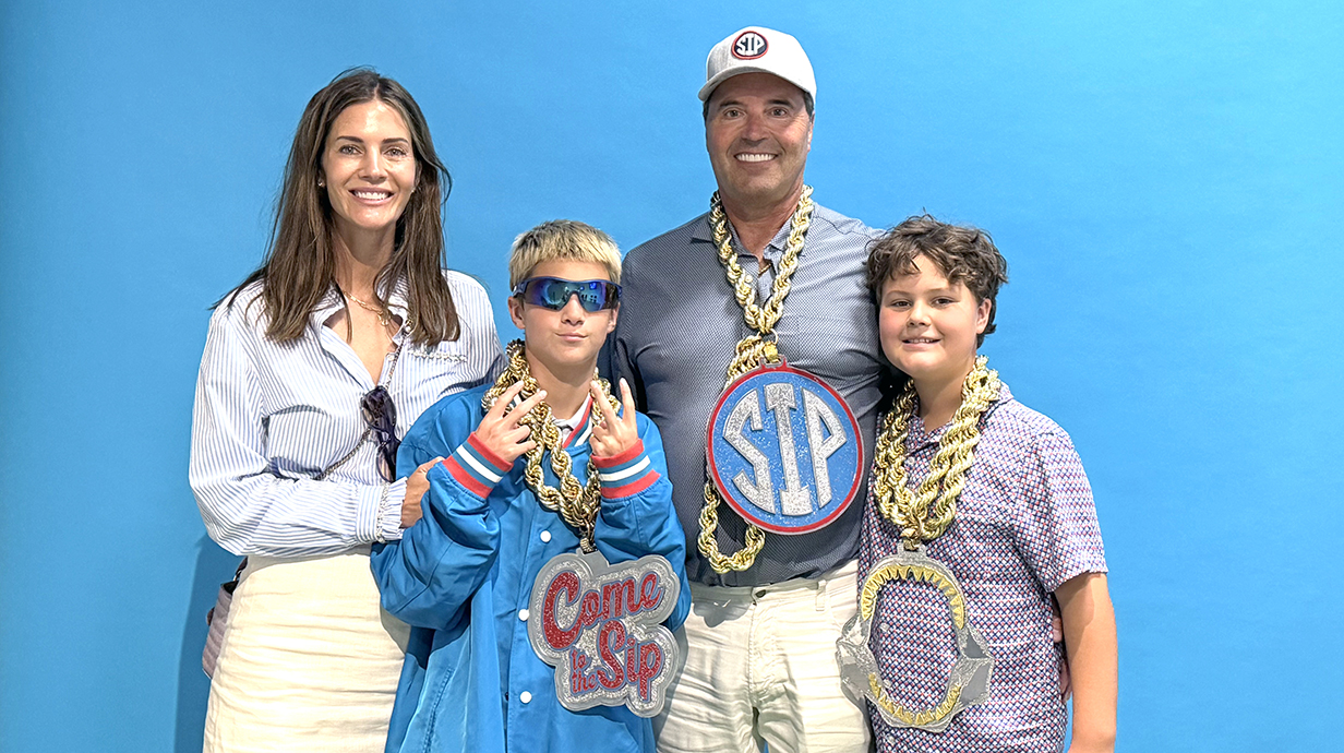 A woman, a man and two boys stand in front of a blue screen while wearing Ole Miss props.