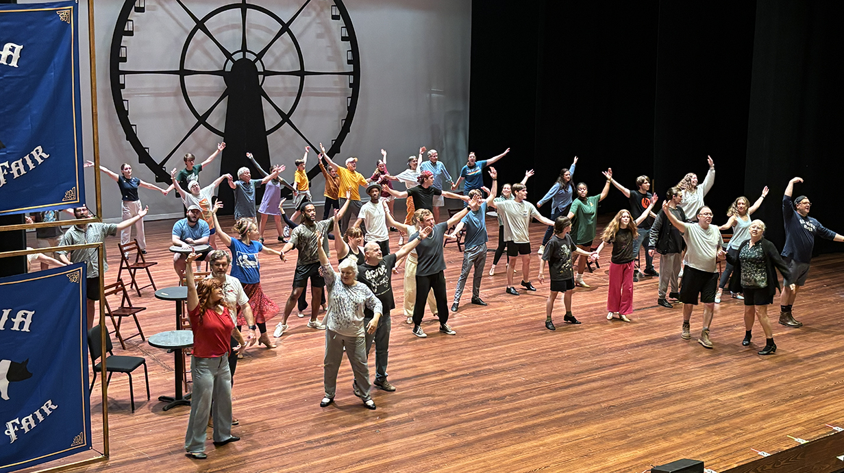 A large group of people rehearses a scene from a play on a stage.