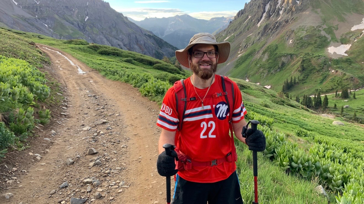 A young man wearing a red Ole Miss jersey and a hat hikes on a trail through mountains.