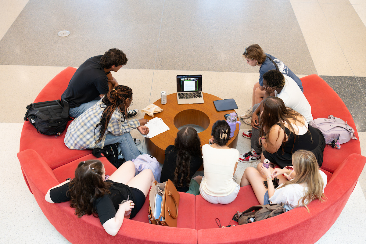 students seated on a circular couch watching an open laptop 
