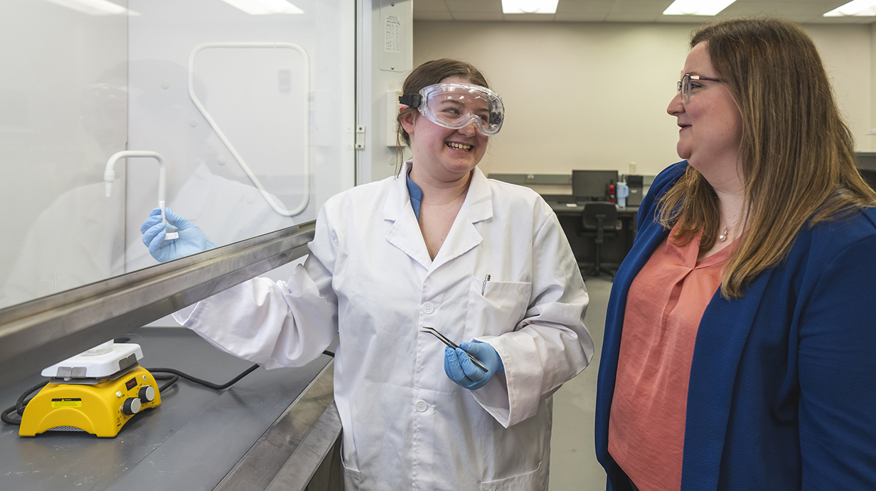 Two women smile standing next to a vented hood in a laboratory.