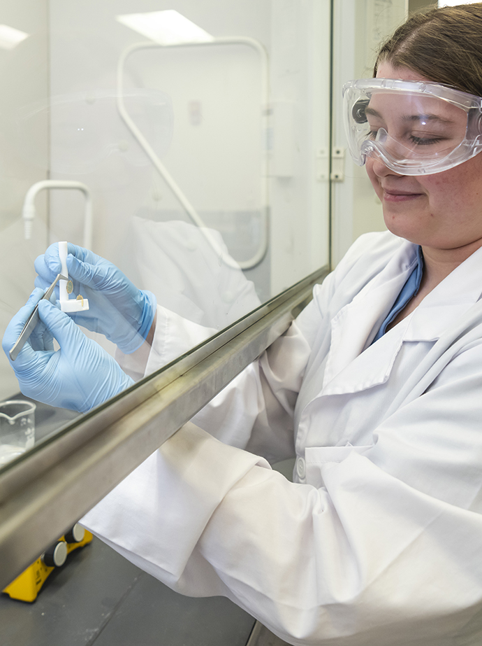 A woman wearing a white lab coat and safety glasses works with a small piece of equipment underneath a vented hood in a lab.