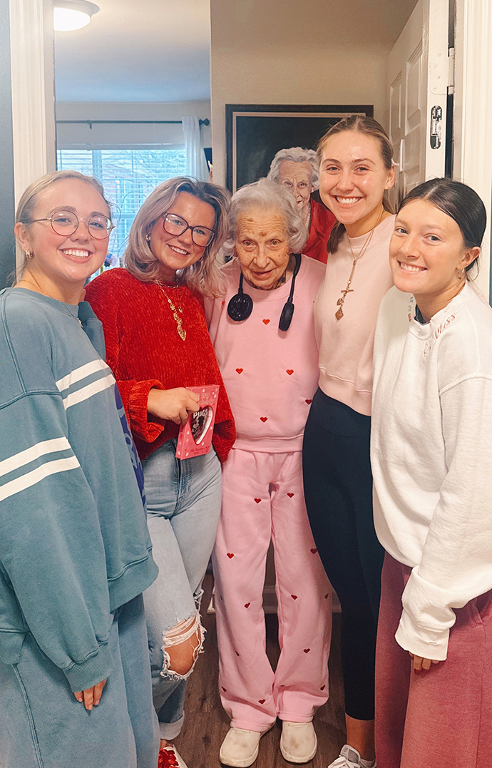 Four young women surround an older woman standing in a hallway.